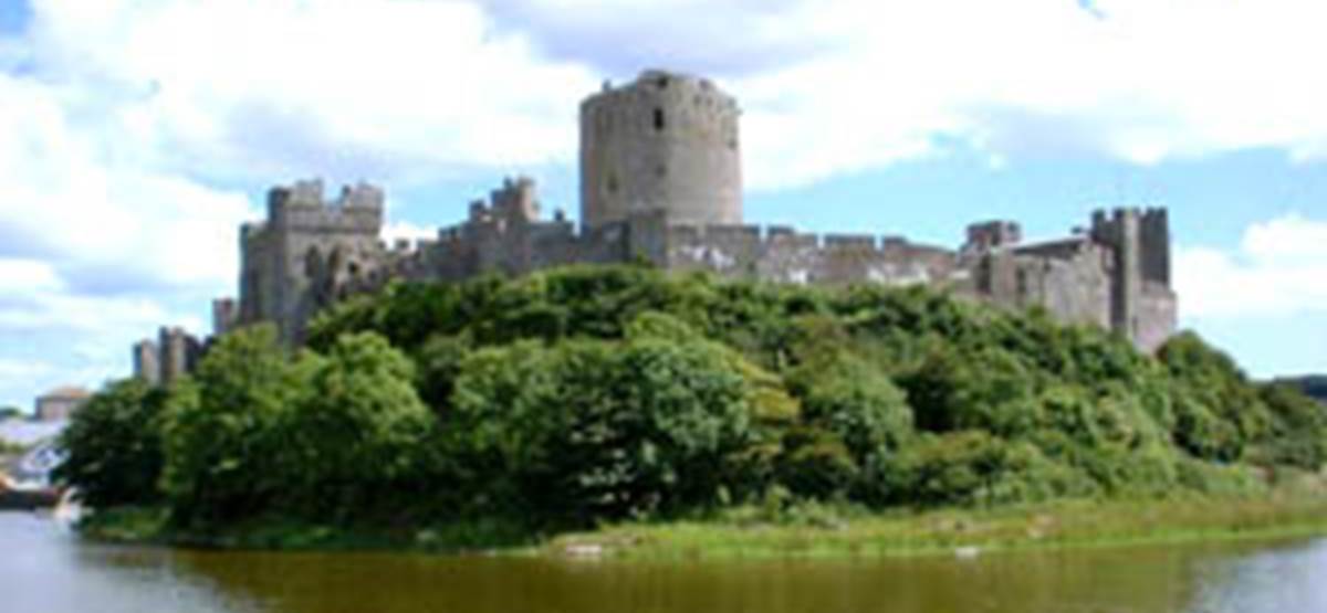 A castle on a hill surrounded by trees with Pembroke Castle in the background
Description automatically generated
