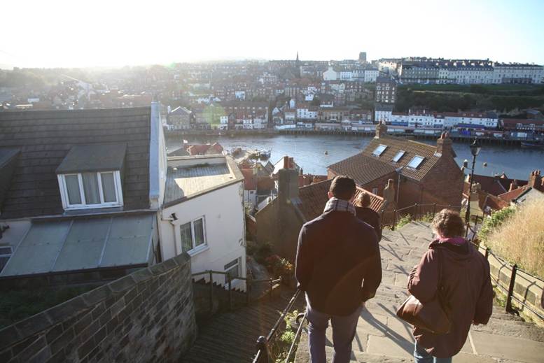 People walking down a hill with buildings and a river
Description automatically generated